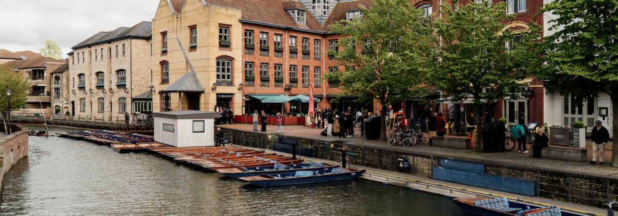 A canal lined with blue punts and floating docks is bordered by historic multi-story brick and stone buildings in Cambridge. People gather on the riverside path, and leafy trees partially shade the scene under a cloudy sky.